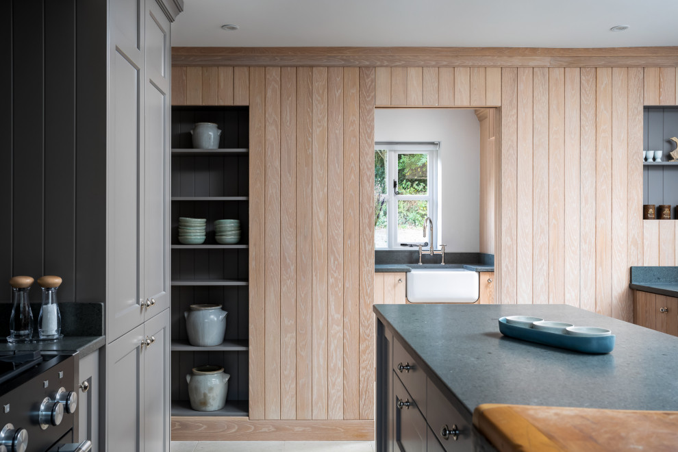 Design ideas for a medium sized beach style galley enclosed kitchen in Gloucestershire with a belfast sink, shaker cabinets, medium wood cabinets, black appliances, an island, beige floors and grey worktops.