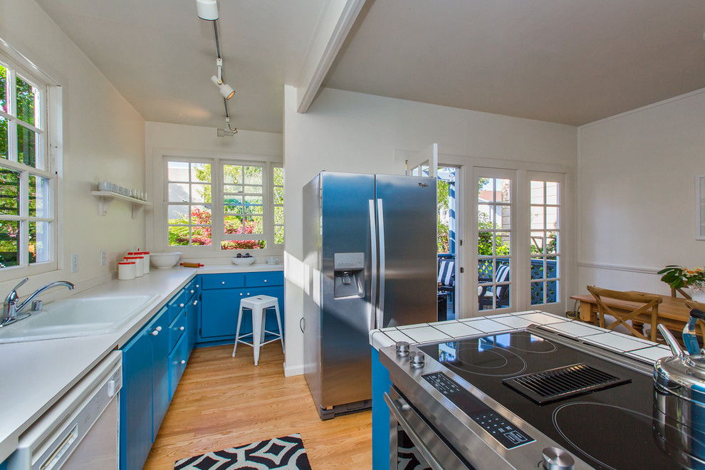 Cette image montre une cuisine américaine parallèle craftsman avec des portes de placard bleues, plan de travail carrelé, un électroménager en acier inoxydable, parquet clair, îlot et un placard à porte plane.