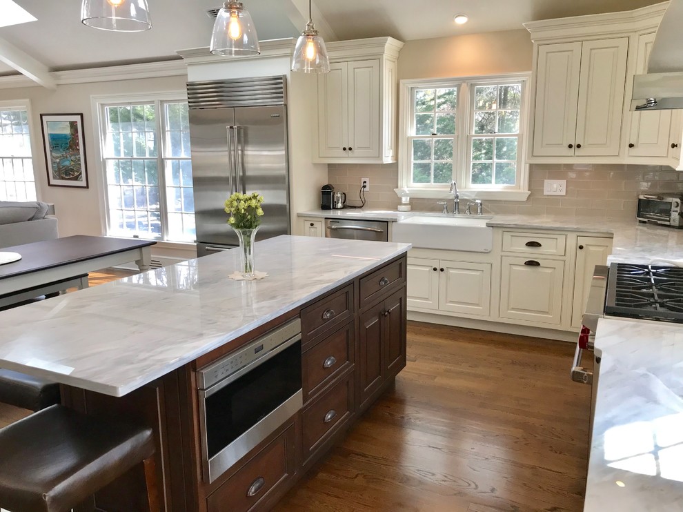 This is an example of an expansive classic l-shaped kitchen/diner in New York with a belfast sink, beaded cabinets, white cabinets, quartz worktops, grey splashback, glass tiled splashback, stainless steel appliances, medium hardwood flooring, an island and brown floors.