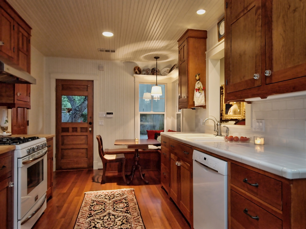 This is an example of a medium sized country galley kitchen/diner in Austin with a double-bowl sink, recessed-panel cabinets, medium wood cabinets, tile countertops, white splashback, ceramic splashback, white appliances and medium hardwood flooring.