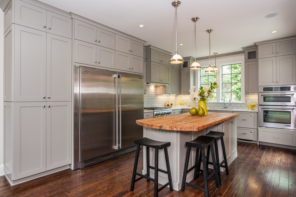 Cette photo montre une cuisine américaine nature en L de taille moyenne avec un placard à porte shaker, des portes de placard grises, une crédence blanche, parquet foncé, îlot, un évier encastré, plan de travail en marbre, une crédence en carreau de verre, un électroménager en acier inoxydable et un sol marron.