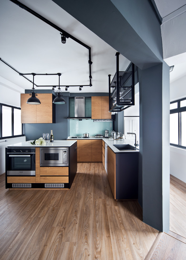 Photo of an industrial l-shaped kitchen in Singapore with flat-panel cabinets, light wood cabinets, glass sheet splashback, stainless steel appliances, light hardwood flooring and an island.
