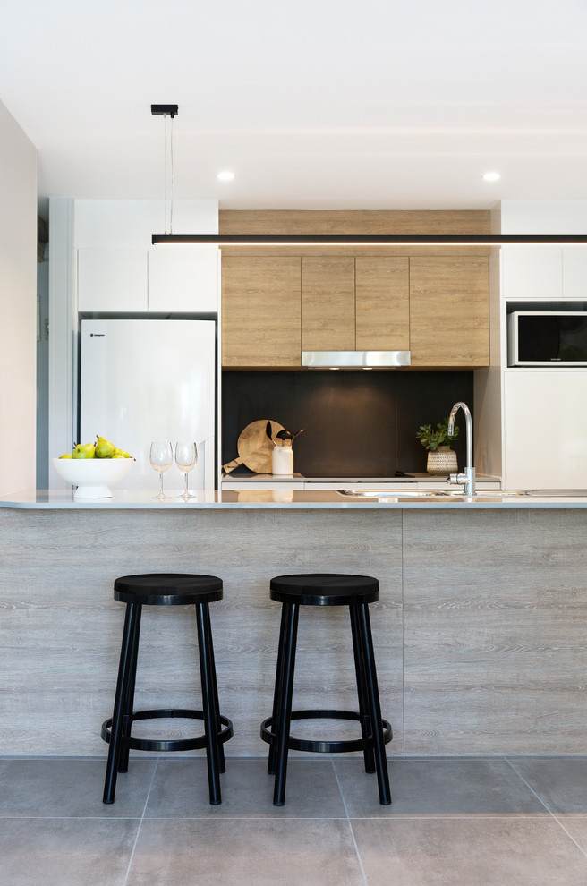 Bild på ett litet funkis grå grått kök, med en undermonterad diskho, släta luckor, skåp i ljust trä, bänkskiva i kvarts, svart stänkskydd, stänkskydd i porslinskakel, vita vitvaror, klinkergolv i porslin, en köksö och grått golv