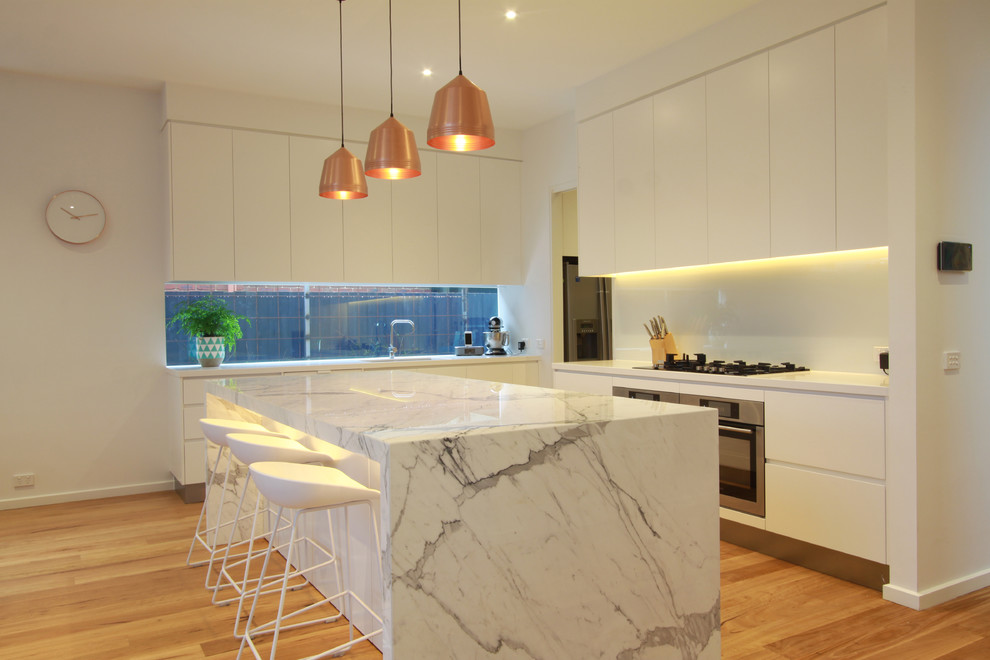 Offene Moderne Küche in L-Form mit Unterbauwaschbecken, flächenbündigen Schrankfronten, weißen Schränken, Marmor-Arbeitsplatte, Küchenrückwand in Weiß, Glasrückwand, hellem Holzboden und Kücheninsel in Melbourne