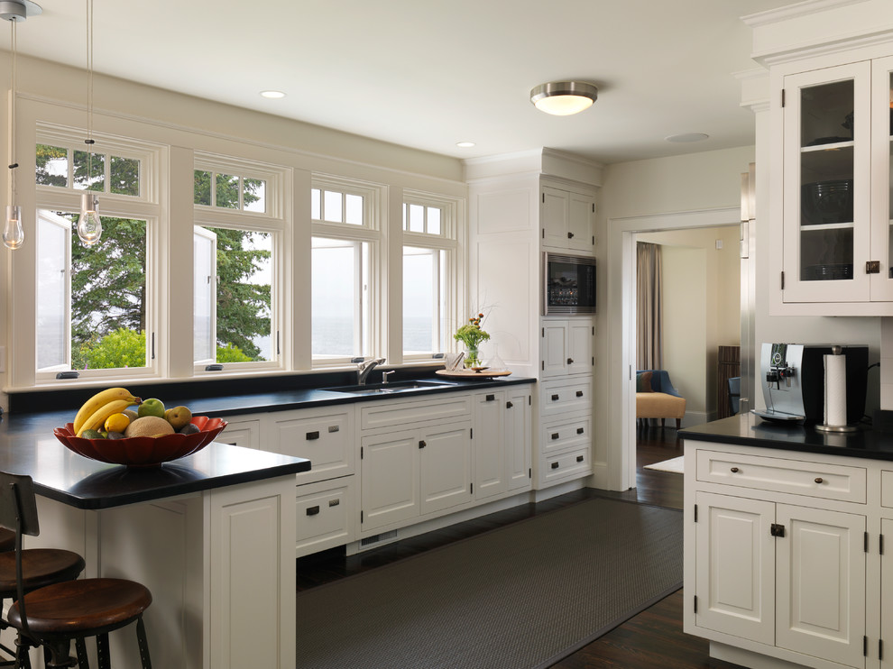 Стильный дизайн: угловая кухня в классическом стиле с врезной мойкой, белыми фасадами, черным фартуком, барной стойкой и окном - последний тренд