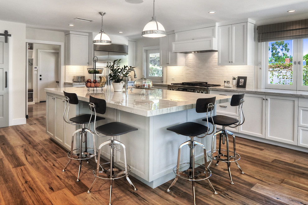 Example of a mid-sized transitional l-shaped medium tone wood floor and brown floor kitchen design in Orange County with white backsplash, an island, an undermount sink, recessed-panel cabinets, white cabinets, stainless steel appliances, subway tile backsplash, quartzite countertops and white countertops