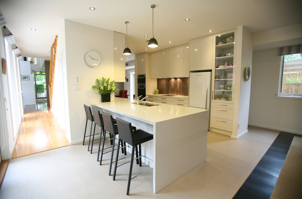 Contemporary galley kitchen in Melbourne with a double-bowl sink, flat-panel cabinets, white cabinets, brown splashback and white appliances.