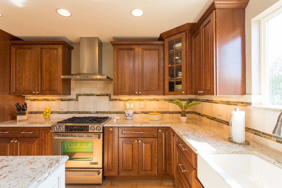 This is an example of a large traditional l-shaped kitchen/diner in Denver with a belfast sink, raised-panel cabinets, medium wood cabinets, granite worktops, beige splashback, stone tiled splashback, stainless steel appliances, an island, medium hardwood flooring and brown floors.