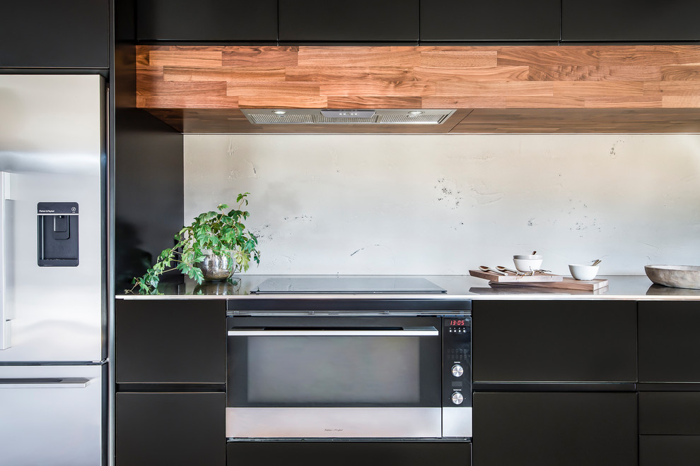Cette photo montre une petite cuisine américaine linéaire moderne avec des portes de placard noires, un plan de travail en inox, une crédence grise et un sol en bois brun.