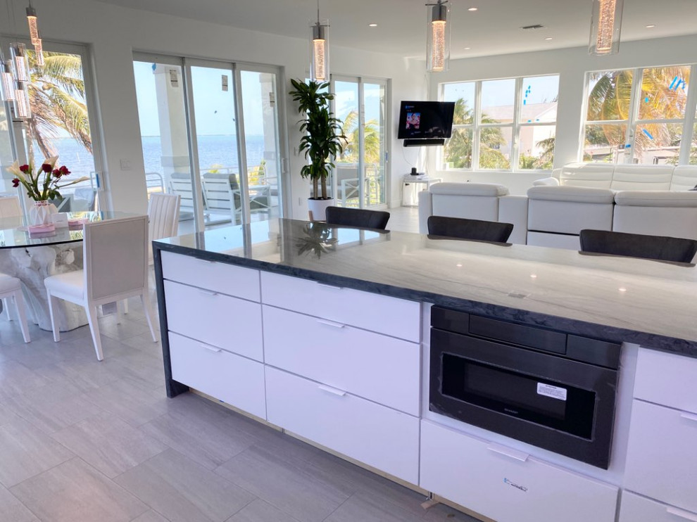 Photo of an expansive contemporary l-shaped kitchen/diner in Miami with a submerged sink, flat-panel cabinets, white cabinets, quartz worktops, white splashback, porcelain splashback, black appliances, an island, grey floors, grey worktops and porcelain flooring.