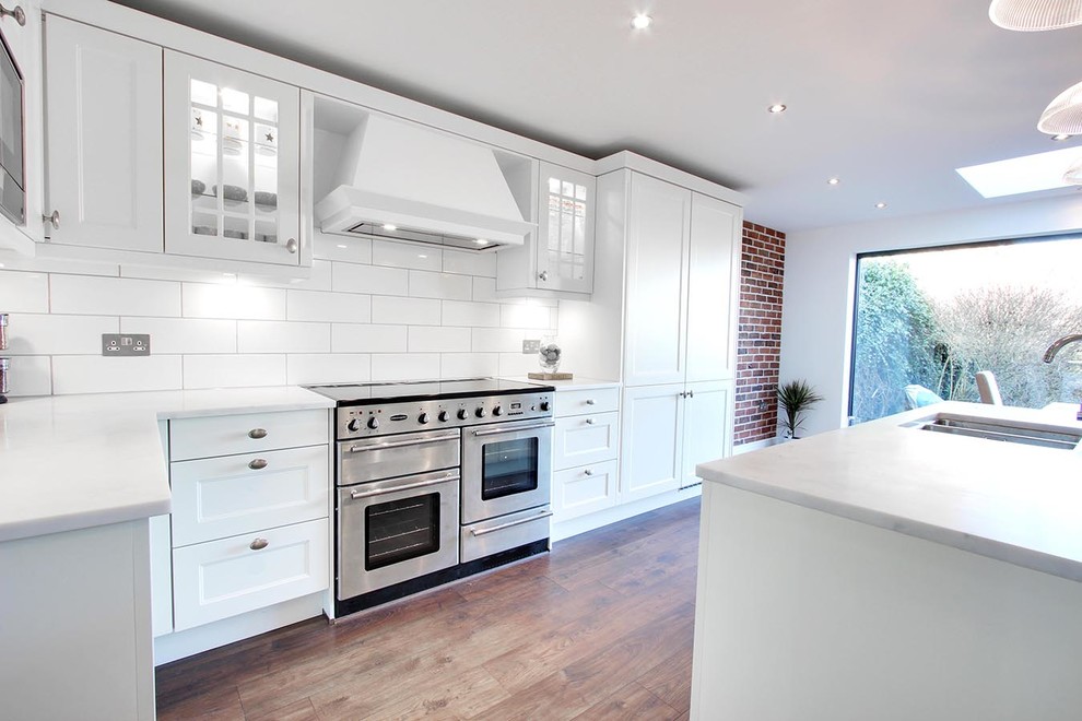 This is an example of a large contemporary galley kitchen/diner in Hertfordshire with a double-bowl sink, shaker cabinets, white cabinets, composite countertops, white splashback, ceramic splashback, stainless steel appliances, painted wood flooring and an island.