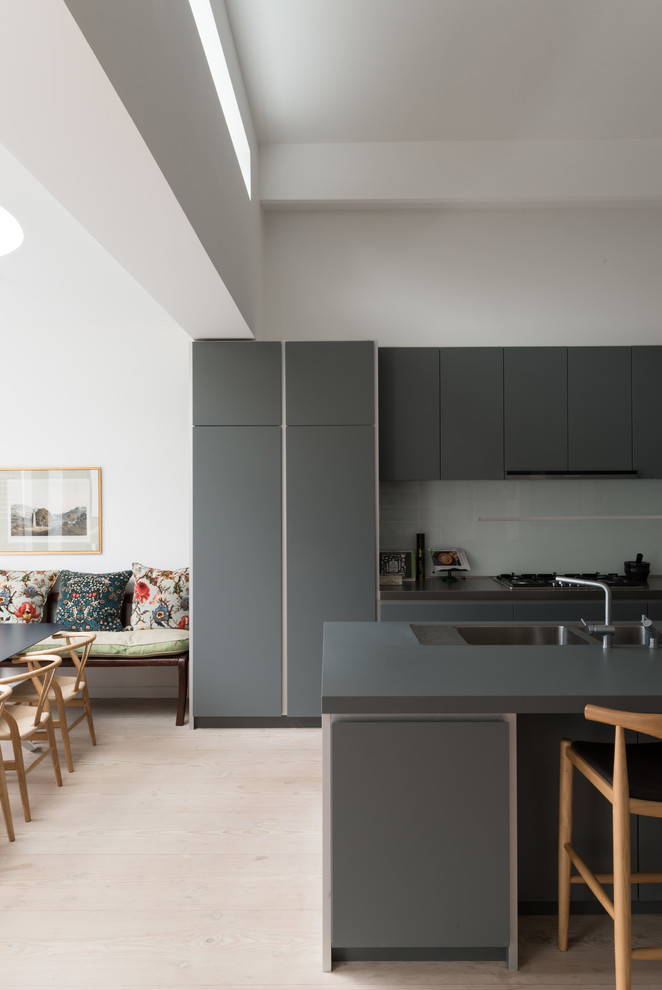 Photo of a medium sized contemporary u-shaped open plan kitchen in London with an integrated sink, flat-panel cabinets, grey cabinets, composite countertops, blue splashback, glass sheet splashback, stainless steel appliances, light hardwood flooring, an island and beige floors.