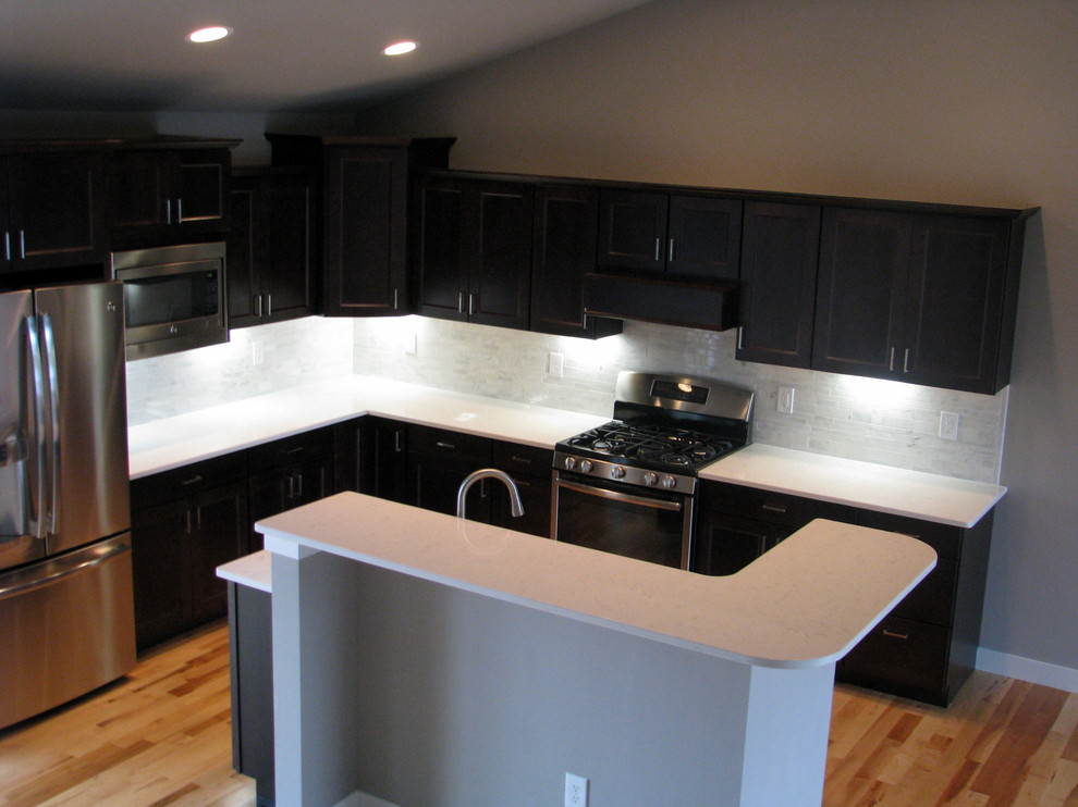 This is an example of a contemporary kitchen in Cedar Rapids with dark wood cabinets, white splashback, stone tiled splashback, stainless steel appliances and light hardwood flooring.