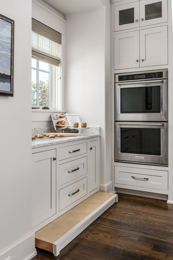 Inredning av ett klassiskt vit vitt kök, med en undermonterad diskho, luckor med profilerade fronter, vita skåp, bänkskiva i kvarts, vitt stänkskydd, stänkskydd i stenkakel, rostfria vitvaror, mörkt trägolv, en köksö och brunt golv