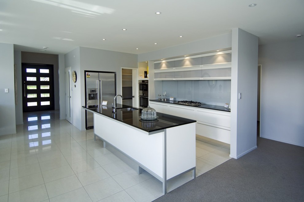 Example of a mid-sized minimalist l-shaped ceramic tile kitchen pantry design in Christchurch with gray backsplash, glass sheet backsplash, an island, a single-bowl sink, flat-panel cabinets, white cabinets, granite countertops and stainless steel appliances