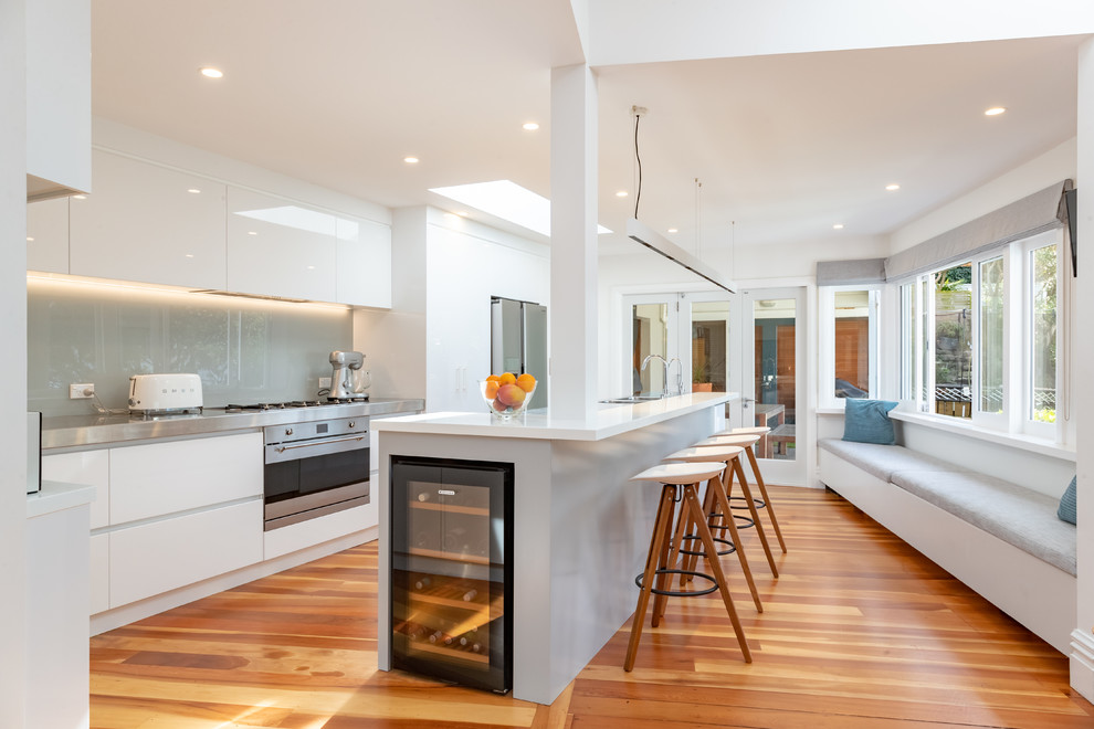 This is an example of a large modern galley kitchen/diner in Wellington with an integrated sink, flat-panel cabinets, white cabinets, stainless steel worktops, grey splashback, glass sheet splashback, stainless steel appliances, medium hardwood flooring, an island and white worktops.
