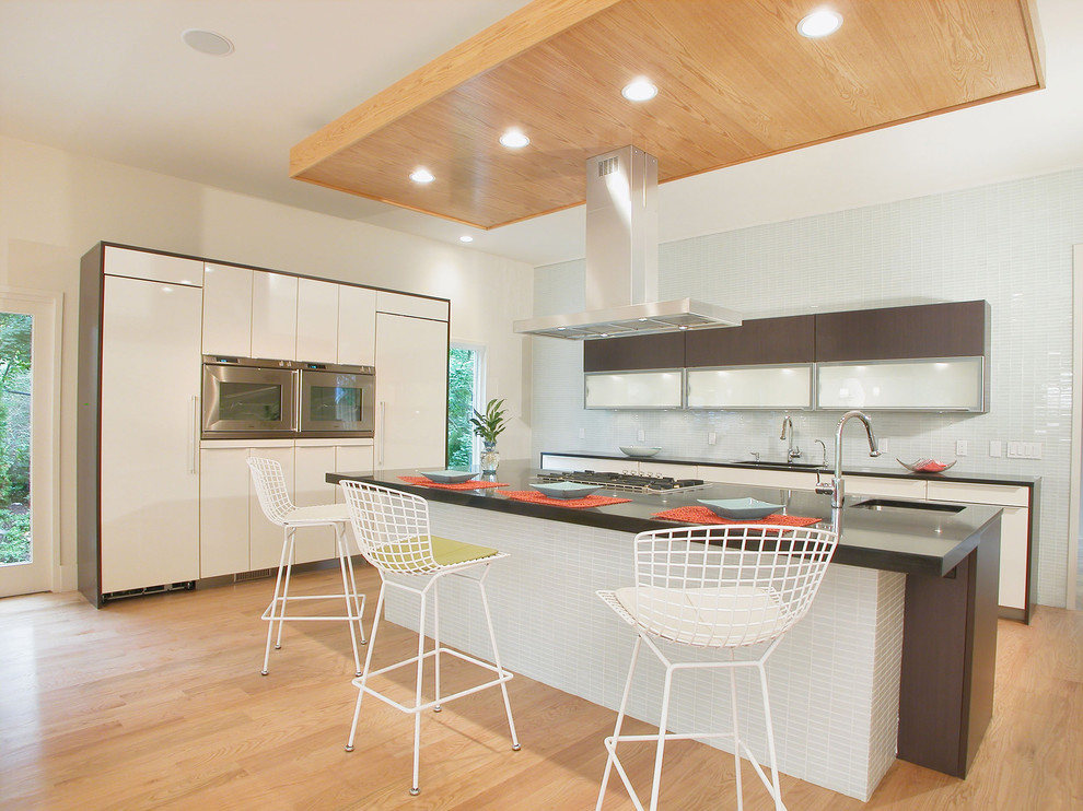 Photo of a contemporary l-shaped kitchen in Philadelphia with flat-panel cabinets, glass tiled splashback, stainless steel appliances, white cabinets, white splashback and light hardwood flooring.