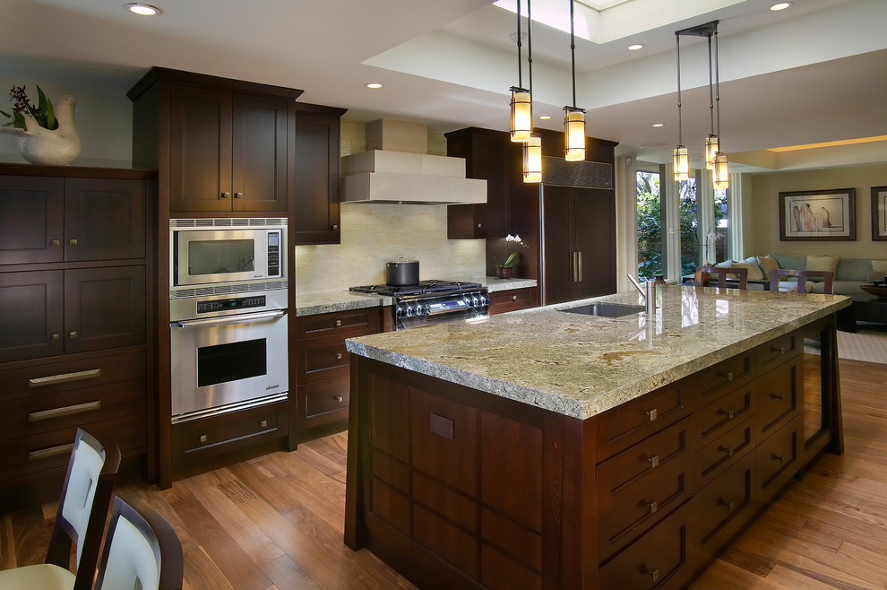 Example of a large trendy l-shaped medium tone wood floor and brown floor open concept kitchen design in San Diego with stainless steel appliances, an undermount sink, shaker cabinets, dark wood cabinets, granite countertops, beige backsplash, stone slab backsplash and an island