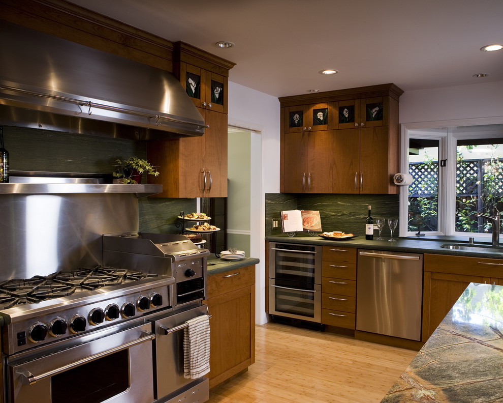 На фото: кухня в современном стиле с техникой из нержавеющей стали, плоскими фасадами, темными деревянными фасадами, зеленым фартуком и фартуком из каменной плиты