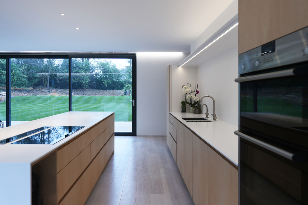Photo of a large contemporary open plan kitchen in Buckinghamshire with an integrated sink, flat-panel cabinets, light wood cabinets, composite countertops, white splashback, integrated appliances, medium hardwood flooring and an island.