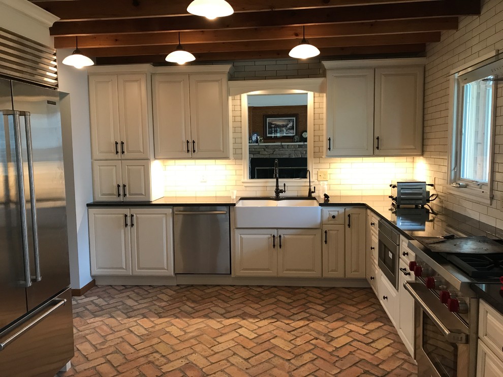 This is an example of a medium sized u-shaped kitchen/diner in Minneapolis with a belfast sink, raised-panel cabinets, white cabinets, granite worktops, white splashback, metro tiled splashback, stainless steel appliances, brick flooring, a breakfast bar, red floors and black worktops.