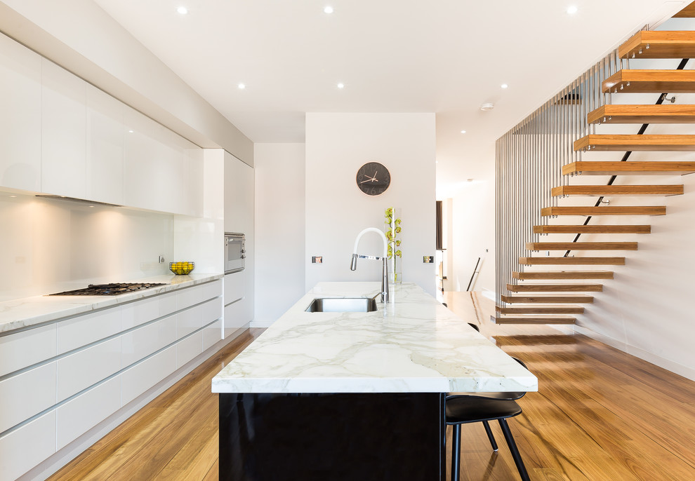 Medium sized contemporary galley kitchen in Melbourne with a submerged sink, white cabinets, white splashback, stainless steel appliances, medium hardwood flooring, an island, brown floors and flat-panel cabinets.