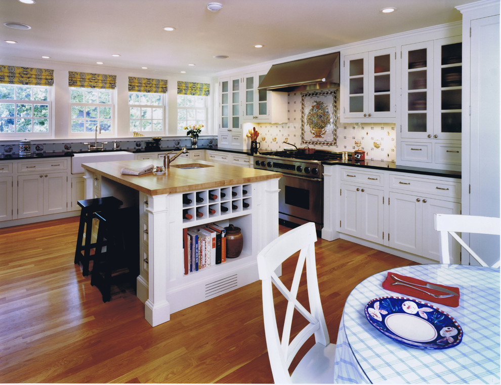 Exemple d'une cuisine chic en L avec un placard à porte vitrée, un électroménager en acier inoxydable, un évier de ferme, des portes de placard blanches, un plan de travail en bois et une crédence multicolore.