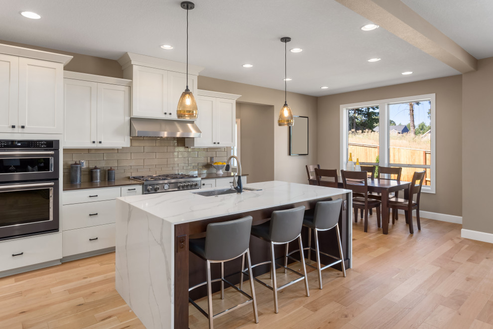 Photo of a medium sized l-shaped kitchen/diner in Los Angeles with a built-in sink, shaker cabinets, white cabinets, engineered stone countertops, beige splashback, metro tiled splashback, stainless steel appliances, light hardwood flooring, an island, beige floors and white worktops.