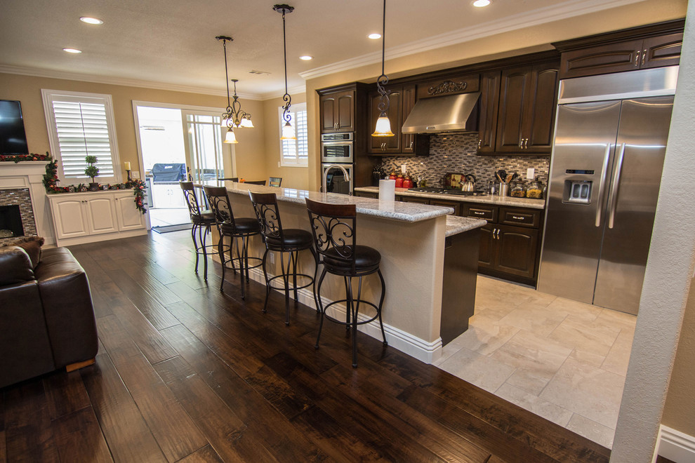 Example of a large transitional travertine floor open concept kitchen design in Orange County with raised-panel cabinets, dark wood cabinets, solid surface countertops, stainless steel appliances and an island