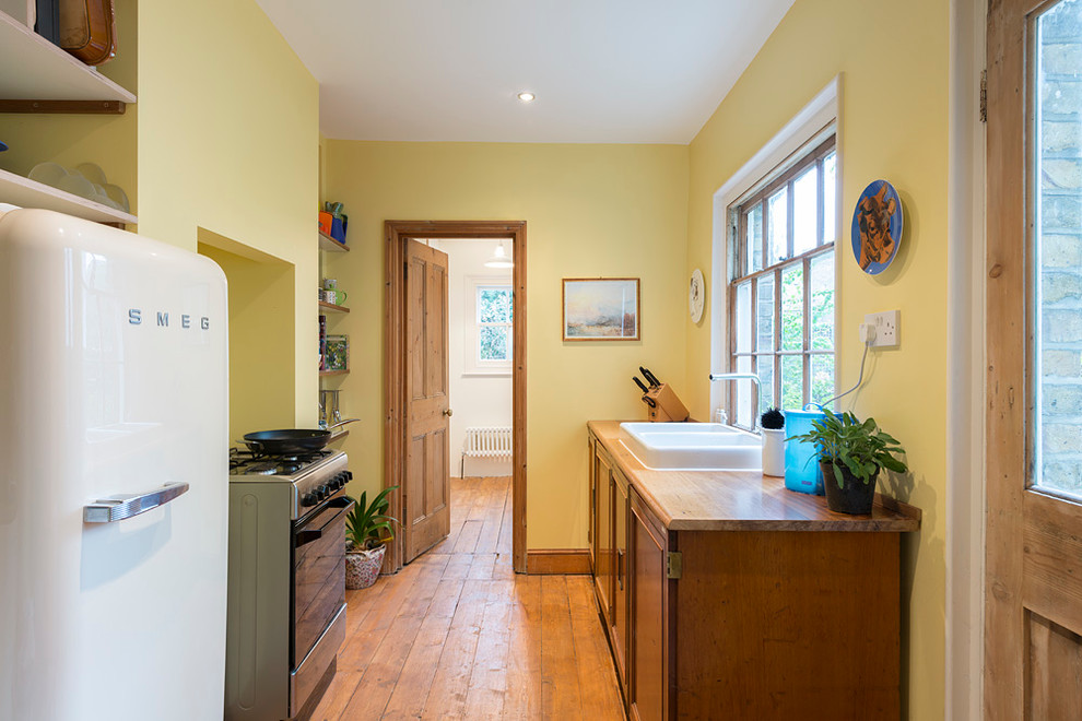 Small classic galley enclosed kitchen in London with a belfast sink, raised-panel cabinets, medium wood cabinets, wood worktops, yellow splashback, stainless steel appliances and medium hardwood flooring.