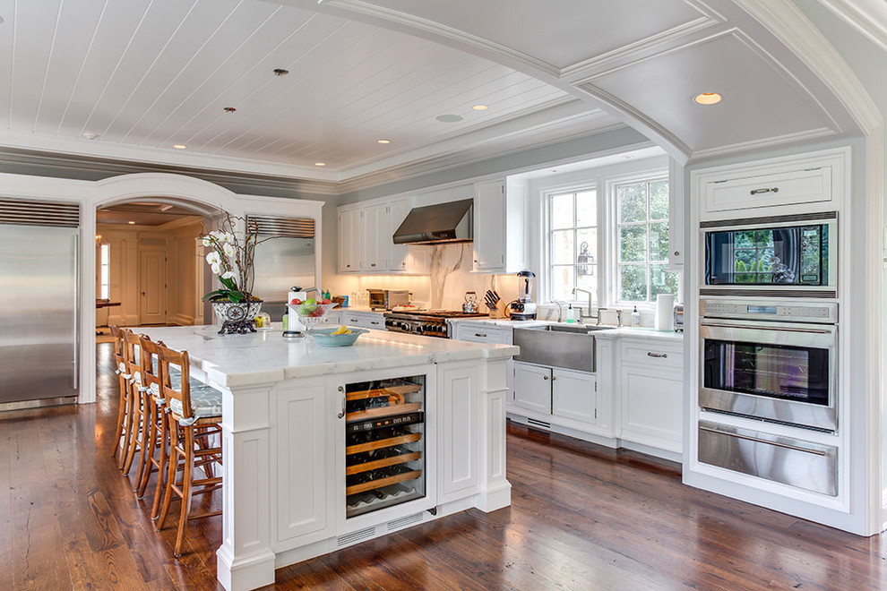 Huge elegant u-shaped brown floor and medium tone wood floor open concept kitchen photo in Other with a farmhouse sink, recessed-panel cabinets, white cabinets, stainless steel appliances, an island, marble countertops, white backsplash and marble backsplash