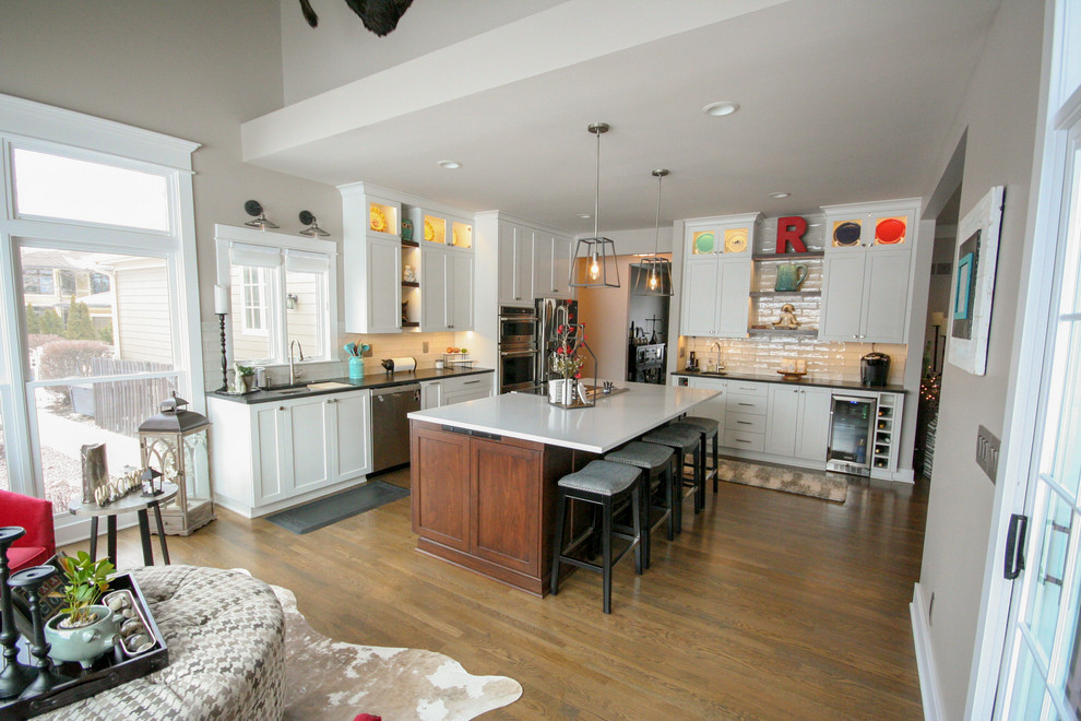 This is an example of a large rural l-shaped open plan kitchen in Kansas City with a submerged sink, shaker cabinets, white cabinets, engineered stone countertops, white splashback, metro tiled splashback, stainless steel appliances, medium hardwood flooring, an island, brown floors and white worktops.