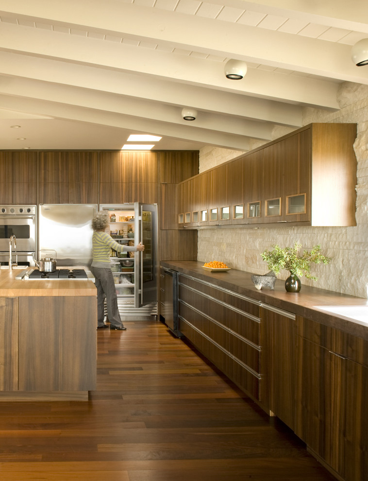 Kitchen - 1950s kitchen idea in Orange County with stainless steel appliances, flat-panel cabinets, dark wood cabinets, wood countertops, beige backsplash and stone tile backsplash