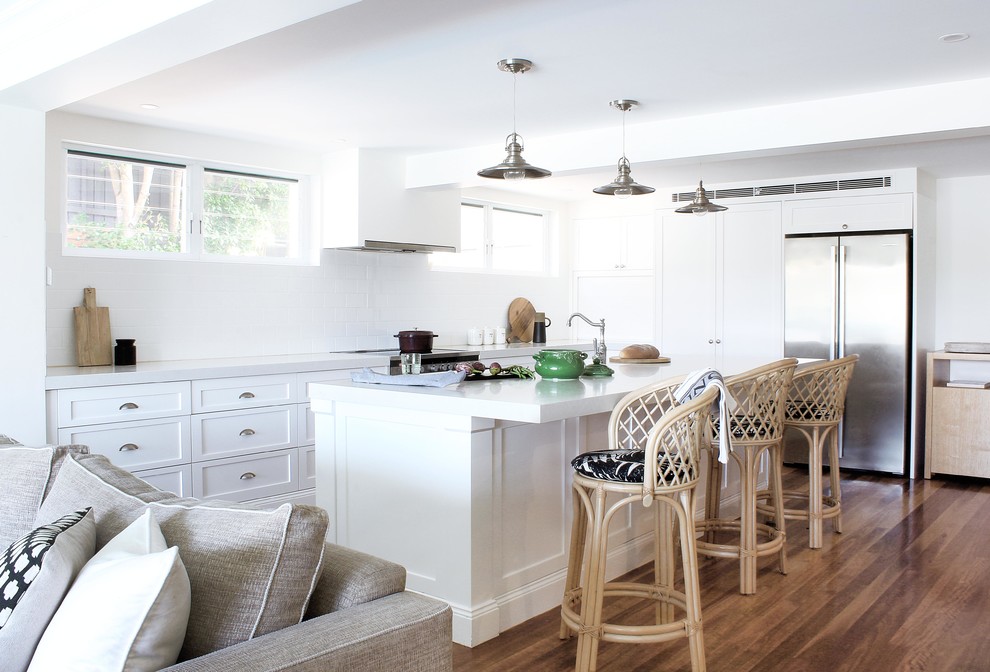 This is an example of a coastal l-shaped open plan kitchen in Sydney with a belfast sink, shaker cabinets, white cabinets, granite worktops, white splashback, ceramic splashback, stainless steel appliances, an island, white worktops, medium hardwood flooring and brown floors.