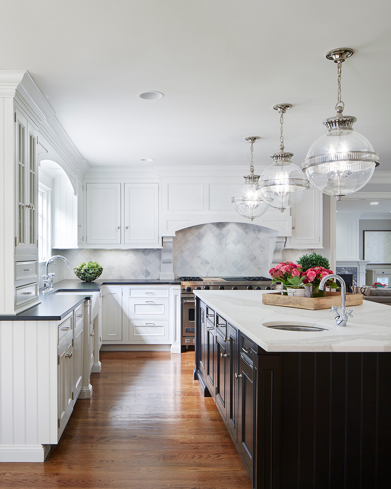 Klassisk inredning av ett kök, med en rustik diskho, luckor med infälld panel, vita skåp, vitt stänkskydd, stänkskydd i marmor, rostfria vitvaror, en köksö och mörkt trägolv