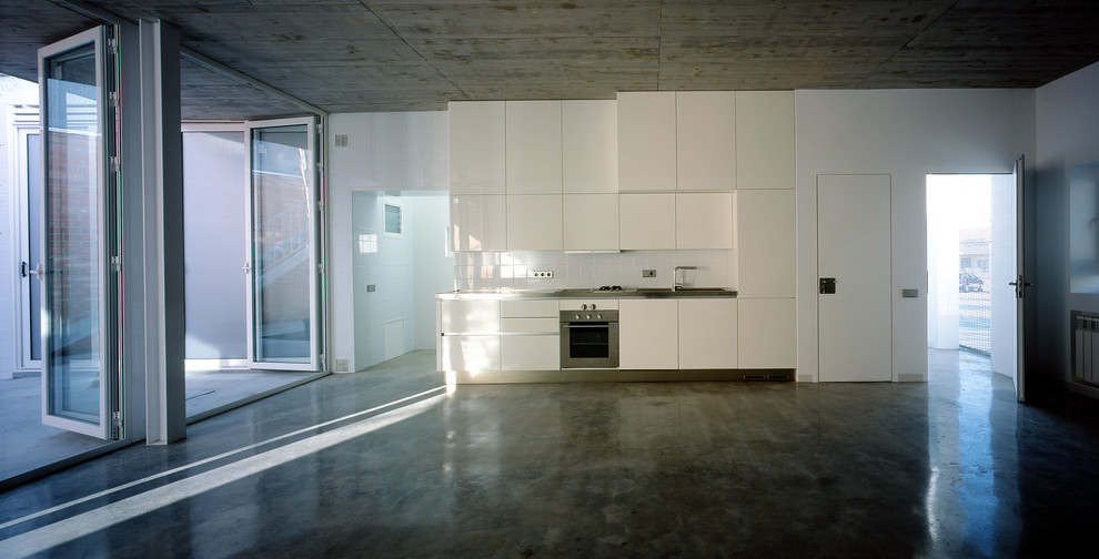 Bild på ett litet funkis linjärt kök med öppen planlösning, med släta luckor, vita skåp, bänkskiva i rostfritt stål, vitt stänkskydd, stänkskydd i porslinskakel, vita vitvaror och betonggolv