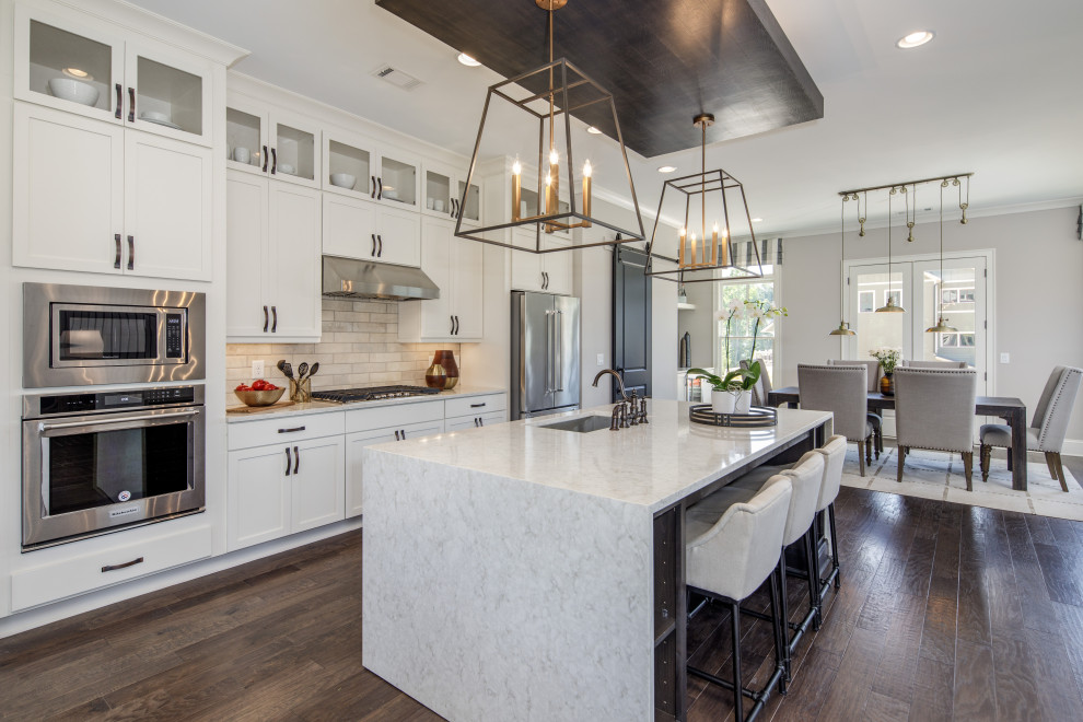 This is an example of a transitional galley kitchen in Atlanta with an undermount sink, shaker cabinets, white cabinets, beige splashback, stainless steel appliances, dark hardwood floors, with island, brown floor and white benchtop.
