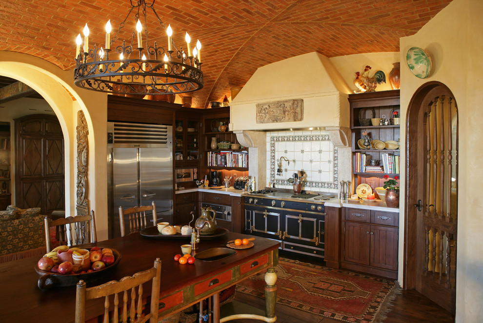 Стильный дизайн: кухня в средиземноморском стиле с обеденным столом, темными деревянными фасадами, разноцветным фартуком и черной техникой - последний тренд