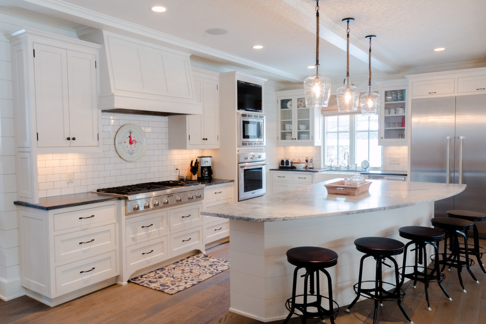 Large traditional l-shaped open plan kitchen in Minneapolis with a belfast sink, beaded cabinets, white cabinets, granite worktops, white splashback, metro tiled splashback, stainless steel appliances, light hardwood flooring and an island.