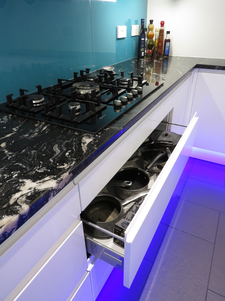 Modern inredning av ett mellanstort l-kök, med granitbänkskiva, en undermonterad diskho, släta luckor, vita skåp, blått stänkskydd, glaspanel som stänkskydd, integrerade vitvaror, klinkergolv i keramik, en köksö och grått golv