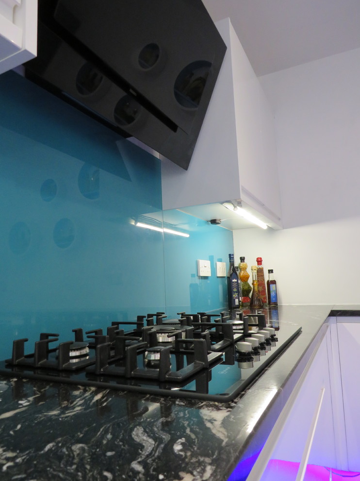 Photo of a medium sized contemporary l-shaped kitchen in Dorset with granite worktops, a submerged sink, flat-panel cabinets, white cabinets, blue splashback, glass sheet splashback, integrated appliances, ceramic flooring, an island and grey floors.