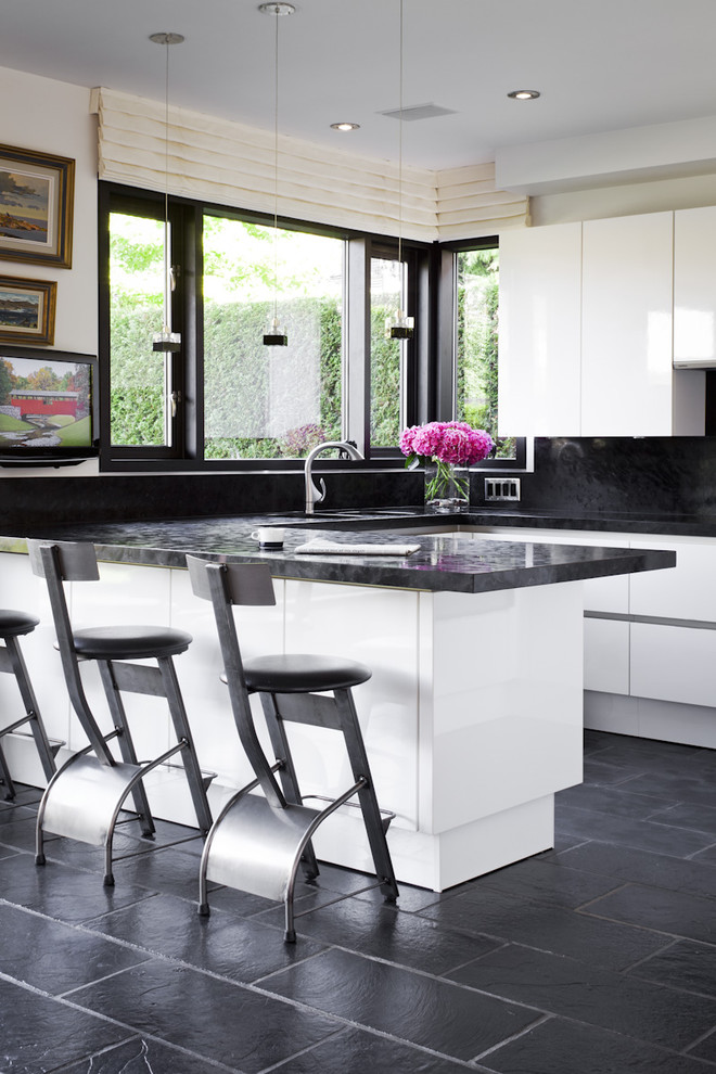 На фото: п-образная кухня в стиле модернизм с плоскими фасадами, белыми фасадами, черным фартуком, черным полом и барной стойкой
