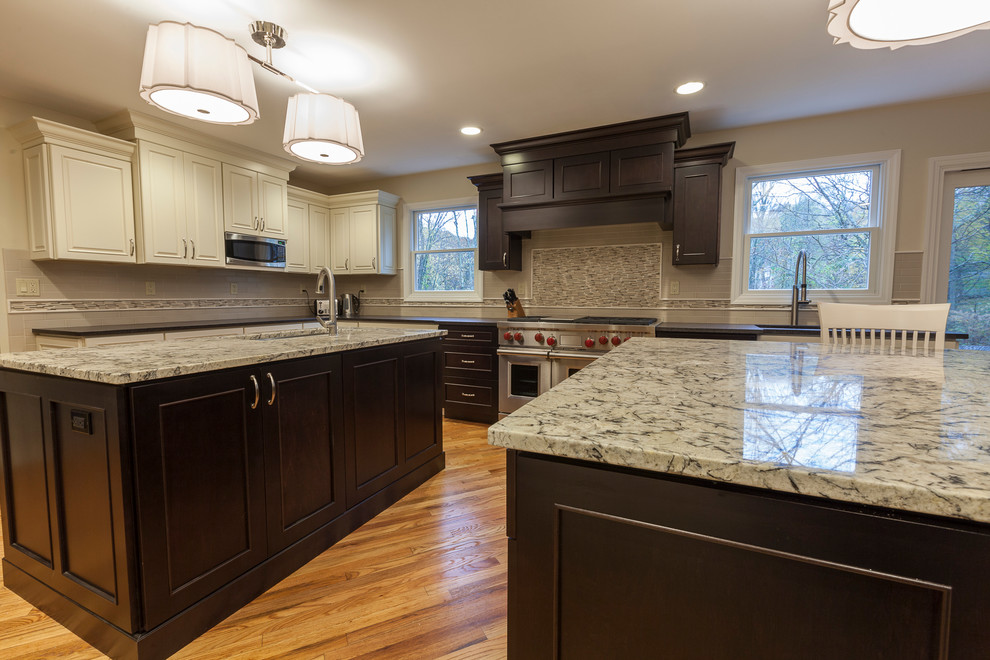 Example of a large transitional l-shaped light wood floor eat-in kitchen design in New York with an undermount sink, raised-panel cabinets, white cabinets, granite countertops, white backsplash, ceramic backsplash, stainless steel appliances and two islands
