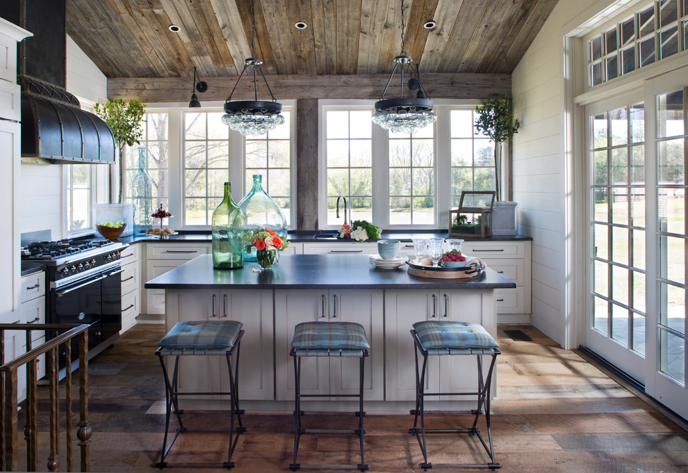 Example of a farmhouse l-shaped medium tone wood floor kitchen design in Austin with an undermount sink, white cabinets, soapstone countertops, black appliances, an island, shaker cabinets, white backsplash and wood backsplash