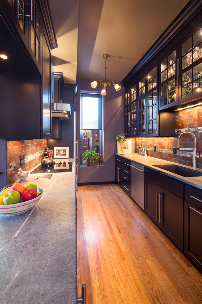 Photo of a small traditional galley enclosed kitchen in Minneapolis with a submerged sink, glass-front cabinets, multi-coloured splashback, stainless steel appliances, medium hardwood flooring, black cabinets, soapstone worktops, porcelain splashback and a breakfast bar.