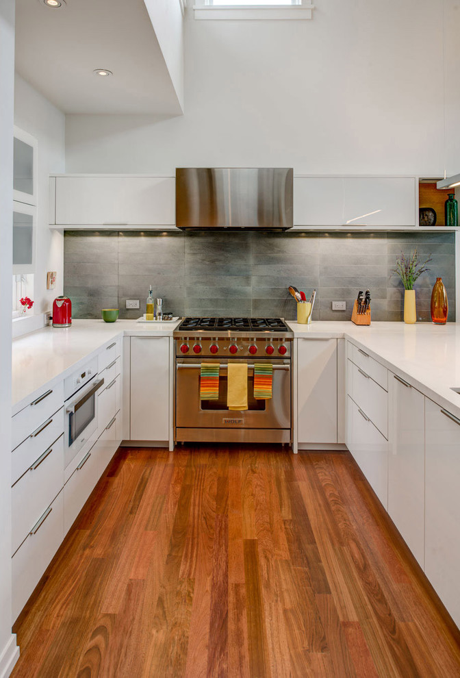 Modern inredning av ett u-kök, med släta luckor, vita skåp, grått stänkskydd, rostfria vitvaror, mellanmörkt trägolv och en halv köksö