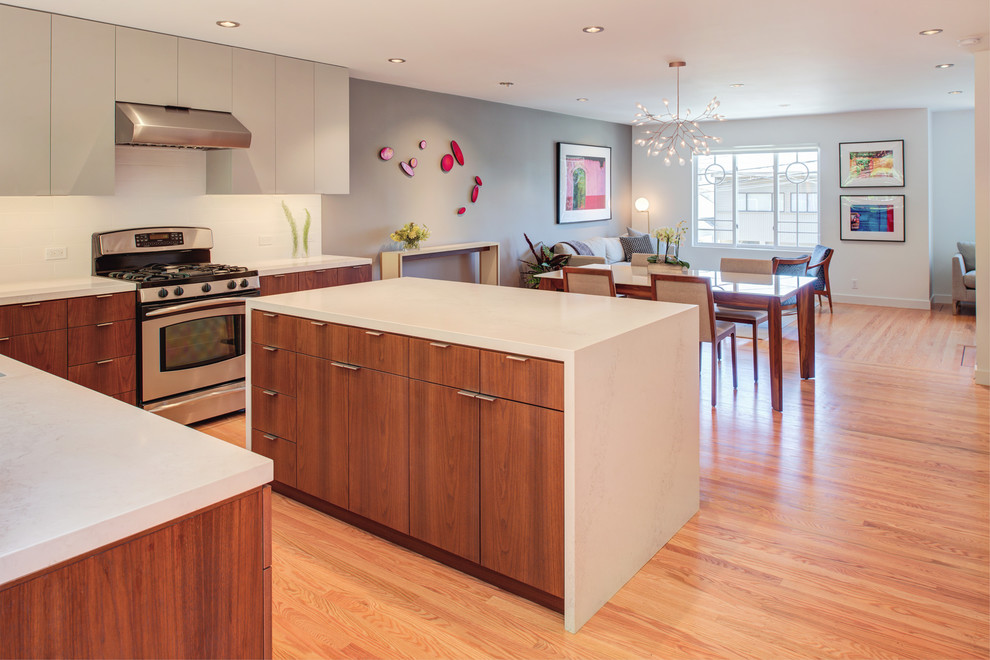 Photo of a small contemporary l-shaped kitchen/diner with a single-bowl sink, flat-panel cabinets, medium wood cabinets, engineered stone countertops, white splashback, stainless steel appliances, light hardwood flooring, an island and white worktops.