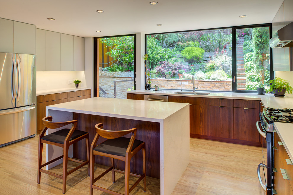 This is an example of a small contemporary u-shaped kitchen in San Francisco with flat-panel cabinets, medium wood cabinets, engineered stone countertops, white splashback, stainless steel appliances, light hardwood flooring, an island, white worktops, a submerged sink and beige floors.