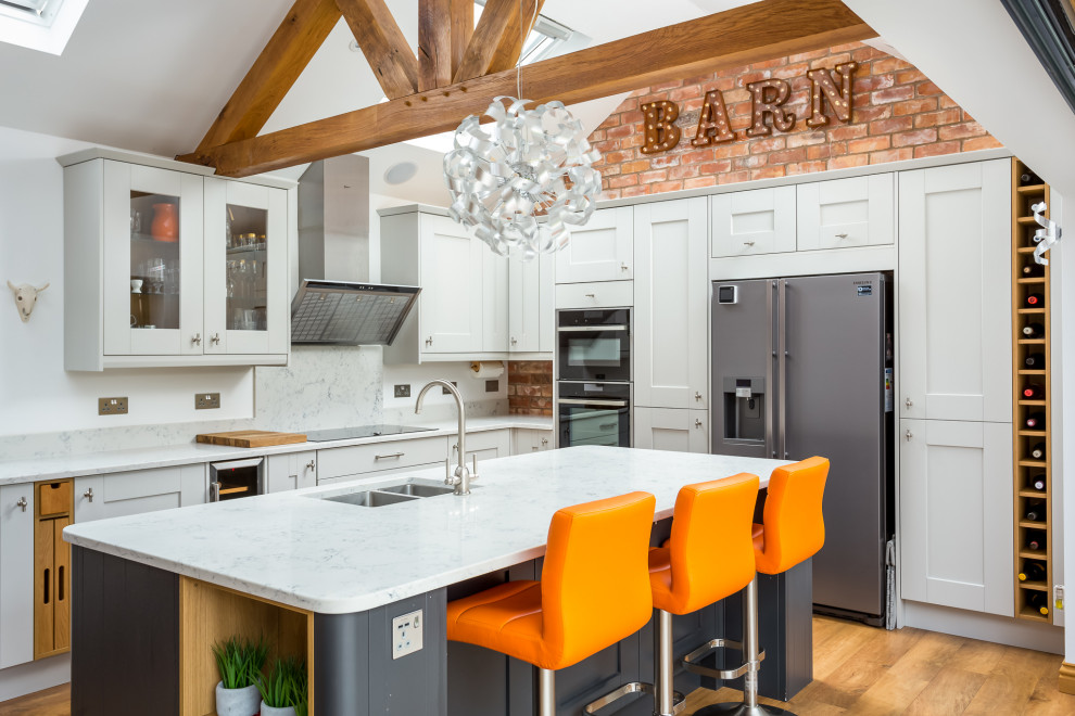 Kitchen - large farmhouse l-shaped brown floor, medium tone wood floor and vaulted ceiling kitchen idea in Gloucestershire with an undermount sink, white cabinets, black appliances, an island, white countertops, shaker cabinets, white backsplash and stone slab backsplash