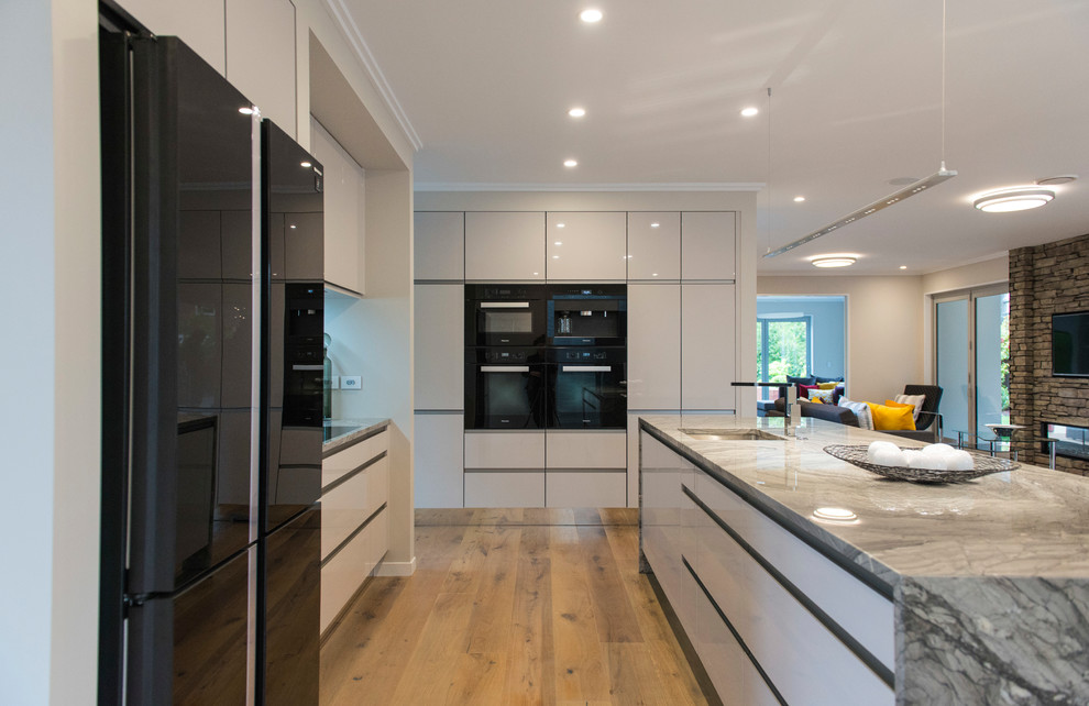 Design ideas for a large contemporary galley kitchen pantry in Christchurch with a submerged sink, white cabinets, marble worktops, multi-coloured splashback, marble splashback, black appliances, laminate floors, an island and brown floors.
